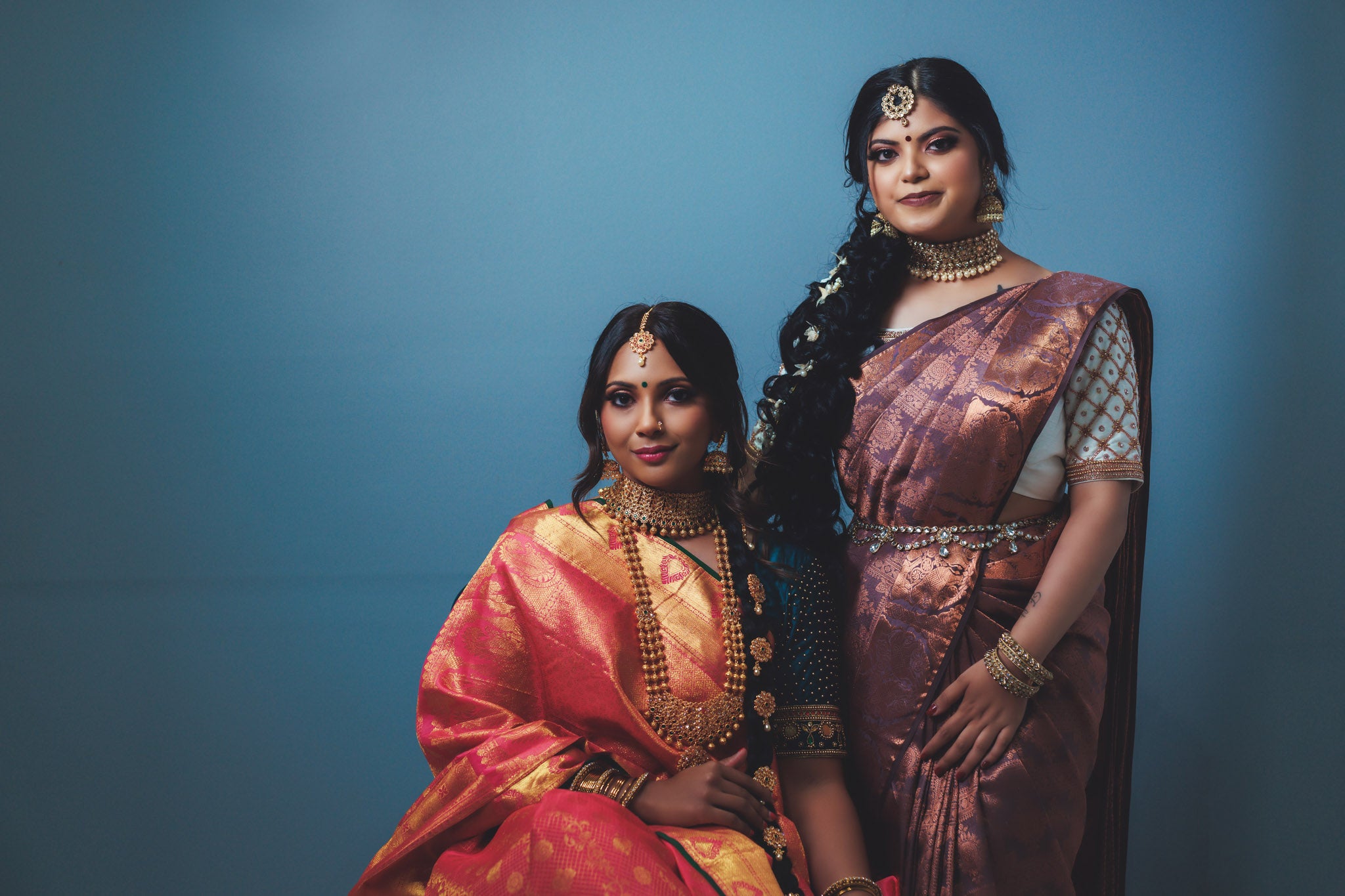 two woman wearing a kanchipuram silk saree. the left woman is wearing a red kanchipuram silk saree with gold zari. the right woman is wearing a maroon kanchipuram silk saree with bronze zari.